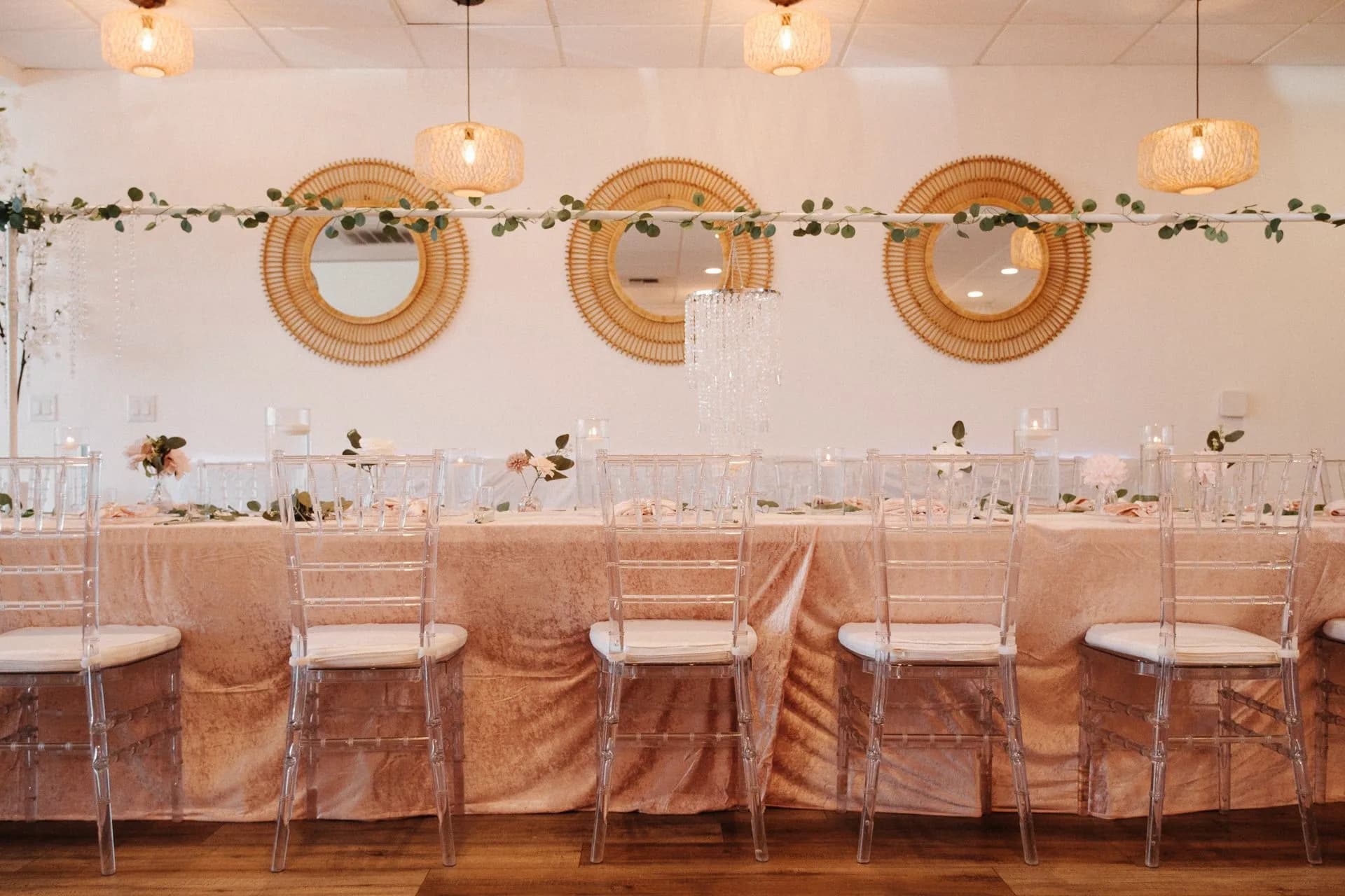 Blush head table with clear chairs, greenery, mirrors, and pendant lights