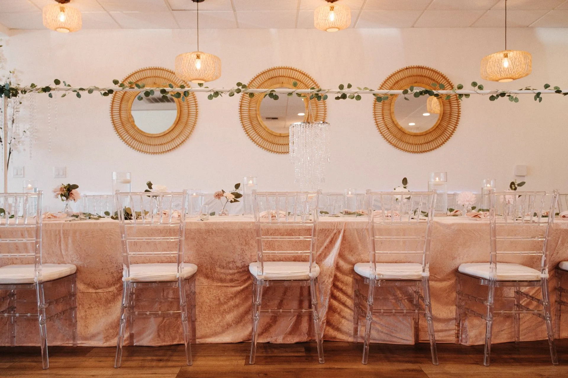 Blush head table with clear chairs, greenery, mirrors, and pendant lights